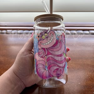 May include: A clear glass tumbler with a bamboo lid and a clear straw. The tumbler features a colorful illustration of the Cheshire Cat from Alice in Wonderland. The cat is pink and purple with a wide grin.