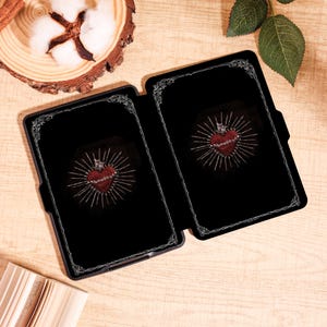 May include: Black tablet case with a decorative silver border. The case is open, revealing a red heart design with a crown and radiating lines on a black background. The design is centered within the case.