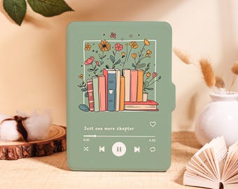 May include: A sage green e-reader case featuring a graphic illustration of a stack of books surrounded by colorful flowers. Below the books, a music player interface displays the text "Just one more chapter" with playback control icons.