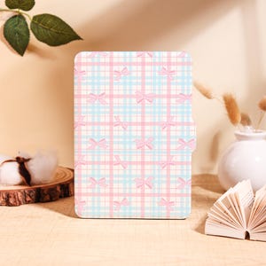 Op de afbeelding: Een rechthoekige hoes met een pastelroze en lichtblauw ruitpatroon, versierd met roze strikken. De hoes is ontworpen voor een tablet of een vergelijkbaar apparaat en biedt bescherming en decoratie.
