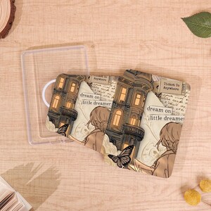 May include: A clear plastic case holds a decorative face mask. The mask features a vintage illustration of a building with lit windows, a woman, and a butterfly. Text on the mask reads "dream on, little dreamer" and "Ticket To Anywhere".