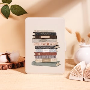 May include: A cream e-reader case with a design of stacked books. The book spines are illustrated with titles such as "Midnights" and "Reputation". The case has a smooth finish and a protective design.