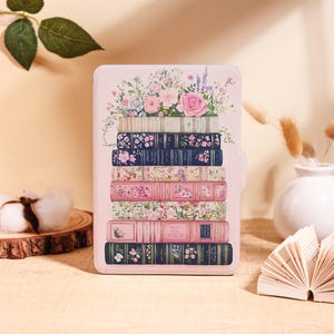 May include: A light pink tablet case with a design of stacked books adorned with floral patterns. The books display a variety of colours and patterns, including pink roses and blue floral prints. The case is designed to protect a tablet.