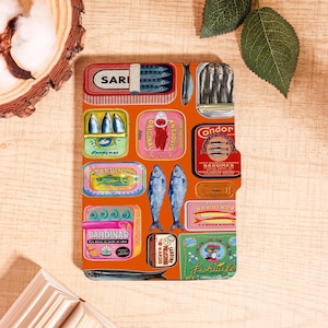 Peut inclure: Un carnet orange avec un motif de boîtes de sardines et d'illustrations de poissons. Les boîtes sont de différentes couleurs et motifs, avec des textes comme "Sari", "Original" et "Condor". Deux poissons bleus sont également représentés.