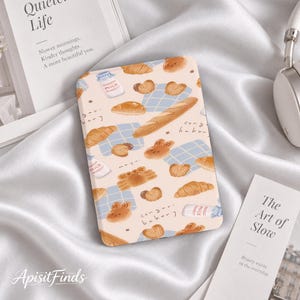 Puede incluir: Una funda rectangular para lector electrónico con fondo rosa claro que presenta un patrón repetitivo de productos horneados marrones, incluyendo croissants, baguettes y panes con forma de animal, junto a botellas de leche y servilletas a cuadros azules.