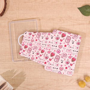 May include: A pink phone case with a Valentine's Day theme. The case features a repeating pattern of hearts, strawberries, keys, and the words "Love" and "Be Mine". A clear plastic case is next to the patterned case.