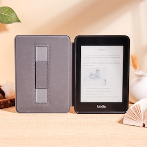 May include: A black Kindle e-reader with a gray cover, open to display text. The cover has a built-in stand. The screen shows text in a foreign language. The Kindle is on a light brown surface.
