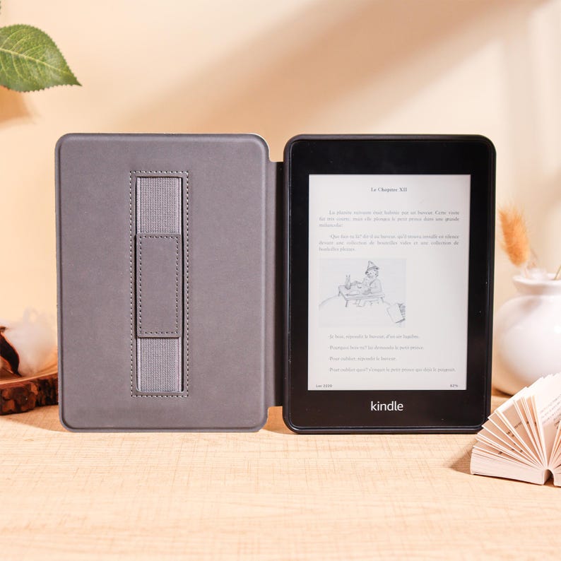 May include: A black Kindle e-reader with a gray cover, open to display text. The cover has a built-in stand. The screen shows text in a foreign language. The Kindle is on a light brown surface.