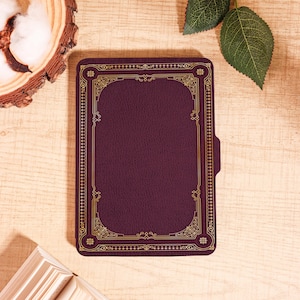 Puede incluir: Una funda rectangular de color burdeos con un borde dorado ornamentado. La funda tiene una superficie texturizada y un bolsillo en el lado derecho. La funda es probablemente para un lector electr&oacute;nico o una tableta. La funda est&aacute; sobre una superficie de madera clara.