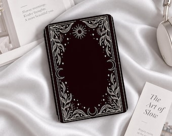 Könnte beinhalten: Eine schwarze E-Reader-Hülle mit einem weißen Linienmuster aus himmlischen Elementen wie Sonne, Mondsicheln, Sternen und Blattzweigen, die in einer dekorativen Umrandung angeordnet sind, platziert auf einem weißen Satinstoff.