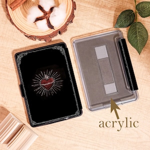 May include: Black e-reader case with a decorative border and a heart design with a crown and thorns. The case is open, revealing a clear acrylic back with a stand. The word "acrylic" is printed on the case.