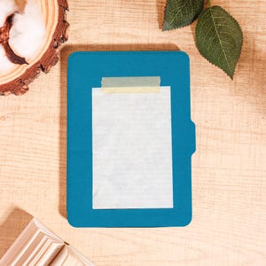 May include: A teal blue folder with a white lined paper taped to it with tan tape. The folder is on a light wood surface, with a small stack of books and a piece of wood with cotton on it.
