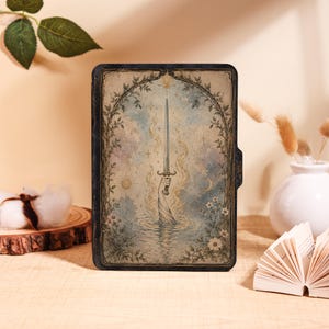 May include: A rectangular e-reader case featuring a detailed illustration of a hand emerging from water holding a sword. The artwork includes a sun, a moon, stars, and floral vines framing the central image in muted blue, gold, and cream tones.