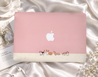 Könnte beinhalten: Eine hellrosa Laptop-Hartschalenhülle mit einem weißen Apple-Logo in der Mitte und einer Reihe von vier kleinen illustrierten Hunden am unteren Rand. Der untere Teil der Hülle ist cremefarben mit dezenten Sprenkeln und bildet einen Kontrast zum rosa Oberteil.