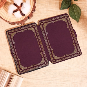 Puede incluir: Una funda de lector electr&oacute;nico burdeos con un dise&ntilde;o de borde dorado ornamentado. La funda est&aacute; abierta, revelando el interior. El dise&ntilde;o se asemeja a una cubierta de libro antiguo. La funda est&aacute; sobre una superficie de madera clara, con una mota de algod&oacute;n y hojas en el fondo.