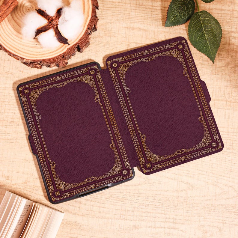 Puede incluir: Una funda de lector electr&oacute;nico burdeos con un dise&ntilde;o de borde dorado ornamentado. La funda est&aacute; abierta, revelando el interior. El dise&ntilde;o se asemeja a una cubierta de libro antiguo. La funda est&aacute; sobre una superficie de madera clara, con una mota de algod&oacute;n y hojas en el fondo.