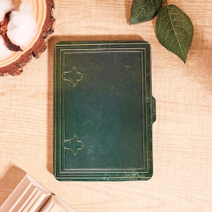 Puede incluir: Un diario de cuero verde oscuro con ribetes dorados y detalles decorativos en las esquinas. El diario es rectangular y tiene una superficie texturizada. La imagen está sobre una superficie de madera clara con elementos decorativos.