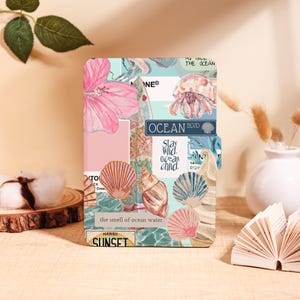 Peut inclure: Un journal décoratif avec un collage d'illustrations sur le thème de l'océan. La couverture présente des coquillages, une fleur d'hibiscus rose et le texte "OCEAN BLVD" et "Stay Wild, ocean child." Le journal a une palette de couleurs bleu clair et rose.