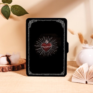 May include: Black tablet case with a red heart design, crown, and radiating lines. The heart has small bead details. The case has a decorative border and a magnetic closure. The background is a light beige.