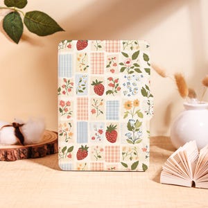 Puede incluir: Un cuaderno color crema con un diseño de patchwork con fresas, flores y cuadros a cuadros. La portada tiene un estampado floral y frutal en rojo, azul y amarillo. El cuaderno está sobre una superficie de madera.