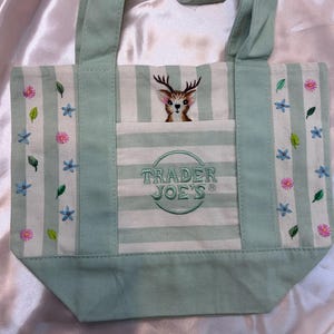 May include: A light green and white striped canvas tote bag featuring an embroidered deer head above a front pocket with the text TRADER JOE'S. The bag is decorated with embroidered pink flowers, blue star-shaped flowers, and green leaves.