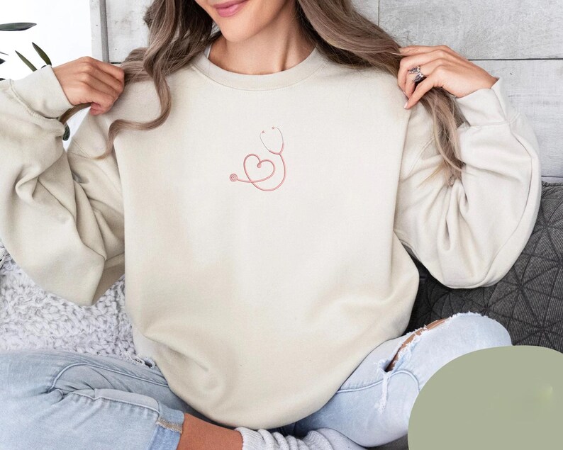 May include: A cream-colored sweatshirt with a pink stethoscope design featuring a heart shape. The sweatshirt is made of a soft, comfortable fabric.