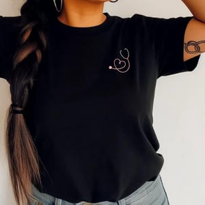 May include: Black t-shirt with a pink stethoscope design on the front. The stethoscope is shaped like a heart.