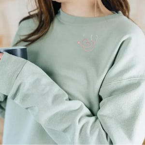 May include: A light green sweatshirt with a pink and white heart and stethoscope design on the front.