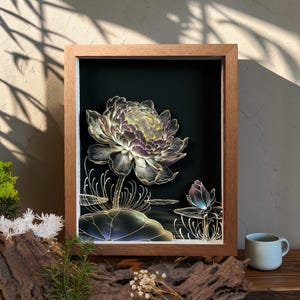 May include: A rectangular wooden shadow box frame containing a layered art piece featuring a large, detailed lotus flower and a smaller bud in shades of purple, white, and gold against a solid black background, with thin, delicate line work.