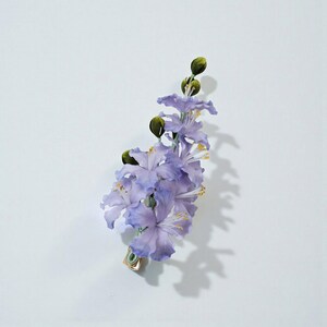 May include: A delicate floral hair clip featuring a cluster of light purple flowers with yellow stamens and green buds. The clip is made of fabric and has a gold-colored metal clasp. The flowers are arranged in a cascading design.