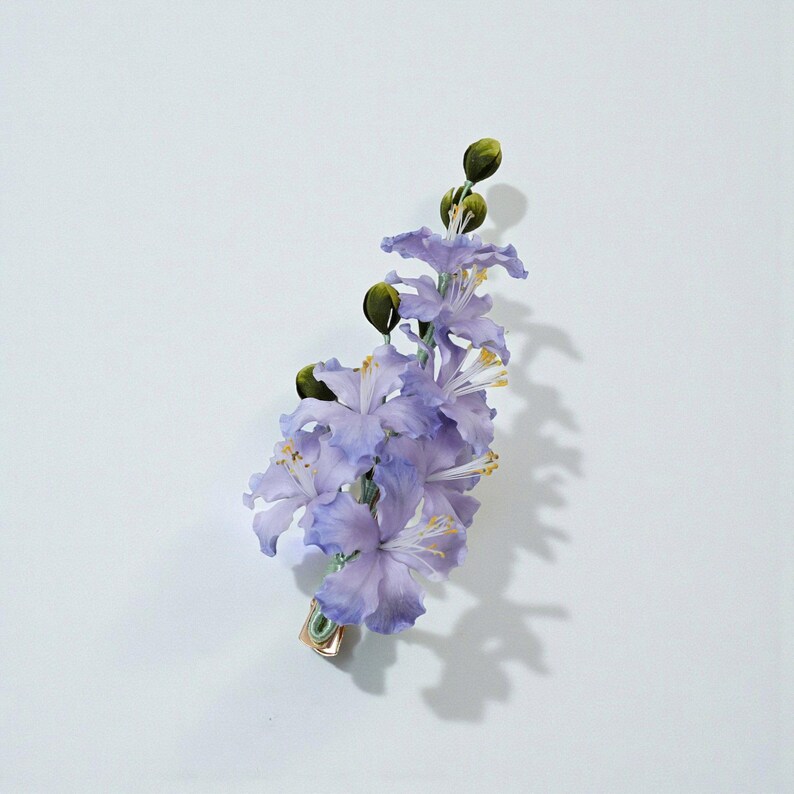May include: A delicate floral hair clip featuring a cluster of light purple flowers with yellow stamens and green buds. The clip is made of fabric and has a gold-colored metal clasp. The flowers are arranged in a cascading design.