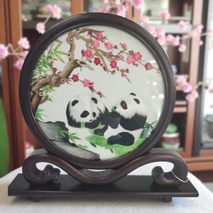 May include: A circular embroidery piece framed in dark wood, featuring two black and white pandas sitting on a green patch. Above them, a brown tree branch extends with bright pink blossoms and green leaves, all set against a white background.