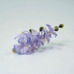 May include: A delicate floral hair clip featuring a cluster of lavender-colored flowers with yellow stamens and green buds. The flowers are attached to a gold-toned metal clip, creating an elegant accessory. The clip is designed to be worn in the hair.