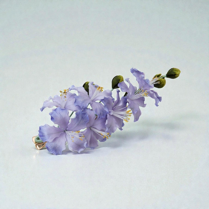 May include: A delicate floral hair clip featuring a cluster of lavender-colored flowers with yellow stamens and green buds. The flowers are attached to a gold-toned metal clip, creating an elegant accessory. The clip is designed to be worn in the hair.