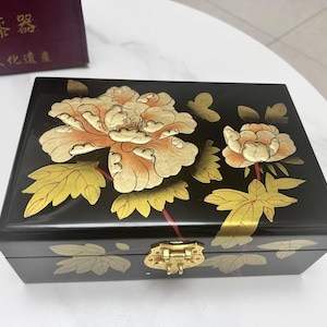May include: Black lacquered wooden box with a gold clasp, adorned with a floral design featuring orange and gold peonies and leaves. The box is rectangular and has a glossy finish, suggesting it is a jewelry or keepsake box.