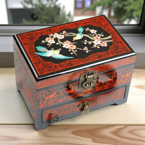 Op de afbeelding: Een zwarte sieradendoos met rode accenten en een gedetailleerd bloemen- en vogelontwerp op het deksel. De doos heeft een lade en een sluiting met een gesp. Het ontwerp is sierlijk en decoratief, met een glanzende afwerking.