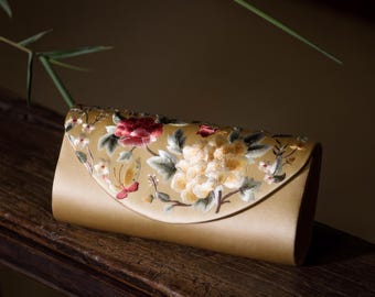 May include: A gold satin clutch with embroidered floral and butterfly details. The embroidery features red, yellow, and green colors. The clutch is rectangular with a curved flap.