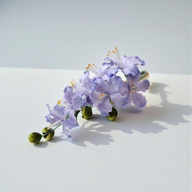 May include: A delicate floral hair accessory featuring light purple flowers with ruffled edges and yellow stamens. The arrangement includes several closed green buds and is set against a white background, casting a soft shadow.