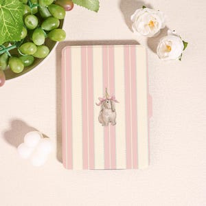 Puede incluir: Un cuaderno a rayas rosas y crema con una ilustración a la acuarela de un conejito con una corona de flores rosas. El cuaderno es rectangular con un borde redondeado. Uvas y flores blancas en el fondo.