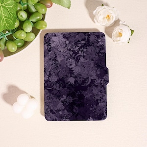 May include: A dark purple tablet case with a floral pattern. The case has a magnetic closure and a textured surface. The design features various shades of purple and gray, creating a layered effect. The case is rectangular and appears to be made of a durable material.