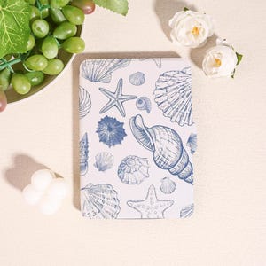May include: A white notebook with a blue seashell and starfish pattern. The notebook is rectangular and features various seashell illustrations. The design includes a starfish, spiral shells, and other sea life. The notebook is a coastal or beach-themed item.