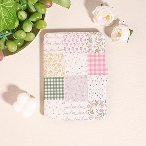 Könnte beinhalten: Ein dekoratives Notizbuch mit Patchwork-Design. Das Notizbuch weist verschiedene Muster auf, darunter rosa Vichy, Blumendrucke und die Worte "love love" in Schreibschrift. Die Farbpalette umfasst Rosa, Grün und Weiß.