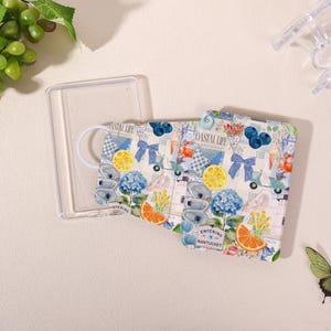 May include: A face mask with a coastal design featuring lemons, blueberries, and hydrangeas. The mask is in a clear plastic case. The text "Entering Nantucket" is visible on the mask.