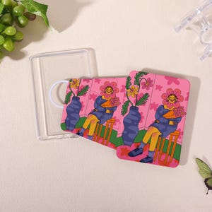 May include: A clear, rectangular phone case with a white loop handle next to two pink, square phone cases. The phone cases feature a floral illustration of a person with a flower face, sitting on a stool, holding flowers, and a vase.