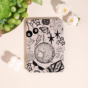 May include: A rectangular e-reader cover featuring a light pink background with a black ink-style print. The design includes a central disco ball, a butterfly, cherries, a martini glass, a slot machine, sunglasses, stars, and a matchbox.