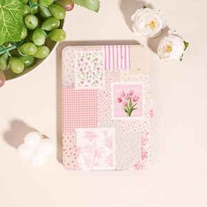 May include: A rectangular notebook with a patchwork design in shades of pink and white. The cover features floral patterns, stripes, and a square with a tulip illustration. The notebook is surrounded by white flowers and a plate of green grapes.