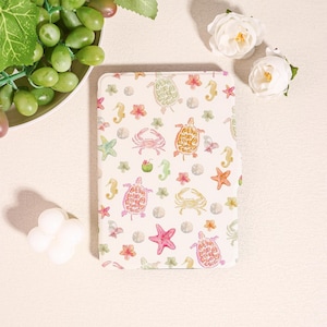 Könnte beinhalten: Ein cremefarbenes Notizbuch mit einem Aquarelldesign von Meeresschildkröten, Seesternen, Seepferdchen und Krabben. Das Notizbuch hat eine weiche, abgerundete Kante und ist mit kleinen rosa Blumen und Sanddollars verziert. Das Gesamtthema ist nautisch.