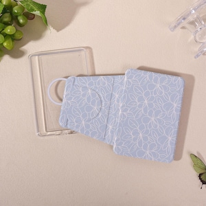 Pode incluir: Uma capa de telemóvel azul-clara com um padrão floral de linhas brancas. A capa apresenta uma moldura de plástico transparente e um painel traseiro dobrável com um recorte circular. Um anel fino está integrado na moldura.