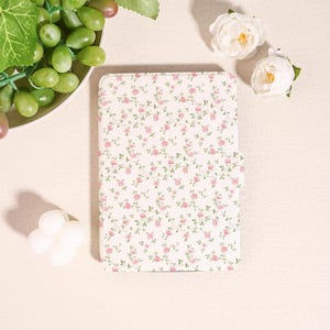 May include: A white tablet case adorned with a pattern of tiny pink roses and green foliage. The case is closed, featuring a magnetic clasp. The design is delicate and floral, suggesting a focus on style and device protection. The case is likely for a tablet.