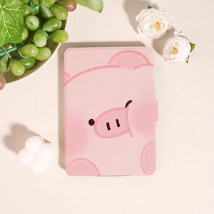 May include: A pink tablet case featuring a cartoon pig face. The case has a magnetic closure and a smooth finish. The pig's face includes a dark brown eye and two black dots for nostrils. The case is designed to protect a tablet.